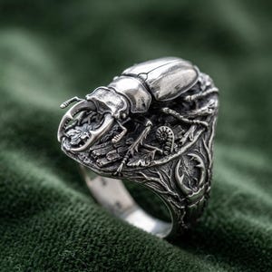 May include: A silver ring featuring a detailed stag beetle sculpture on the top, with textured branch and leaf motifs wrapping around the band. The metal has a polished finish on the beetle and an oxidised finish in the recessed areas.