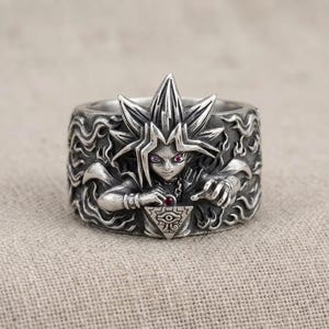 Puede incluir: Un anillo de metal plateado con un relieve detallado de un personaje de pelo puntiagudo que sostiene un colgante triangular con un símbolo de ojo. La banda está decorada con patrones de llamas y pequeños detalles en color rojo.