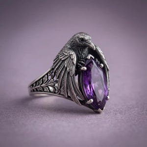 May include: Silver-toned ring featuring a detailed raven design holding a large, teardrop-shaped purple gemstone. The ring has intricate floral patterns on the band, creating a gothic or fantasy aesthetic. The gemstone is approximately 1 inch in length.