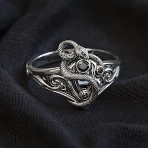 Könnte beinhalten: Ein silberner Ring mit einem detaillierten Schlangendesign, das sich um einen zentralen schwarzen Edelstein windet. Der Ring hat kunstvolle Schnörkel und ist mit kleineren roten Edelsteinen akzentuiert, was eine gotische oder Fantasy-Ästhetik erzeugt. Der Ring wird auf dunklem Stoff präsentiert.