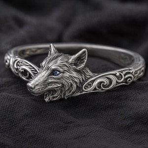 May include: A silver-colored bracelet featuring a detailed wolf head with a blue gemstone eye. The bracelet has an intricate design with swirling patterns on the band. The wolf's fur is meticulously rendered, giving it a realistic appearance.