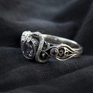May include: A silver ring with a snake design, encircling a large, oval purple gemstone. The ring has detailed patterns and two smaller purple gems. The ring is set against a dark grey backdrop.