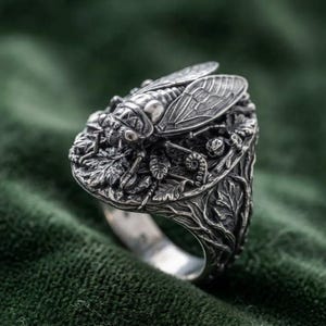 May include: A silver ring featuring a detailed cicada insect resting on a bed of textured leaves and vines. The metal has an oxidized finish that highlights the intricate veins on the insect wings and the organic patterns of the foliage.