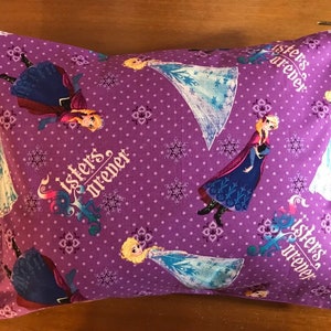 May include: A rectangular cushion with a purple background featuring a pattern of cartoon characters and the text "Sisters Forever." The characters are depicted in various poses, with snowflakes and floral designs scattered across the fabric.