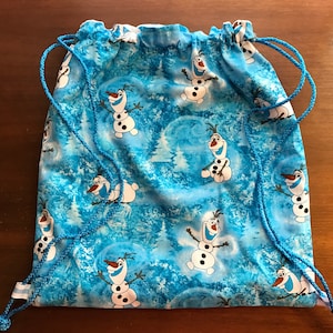 May include: A blue drawstring backpack featuring a repeating pattern of the snowman Olaf from the movie Frozen. The background has a winter forest scene. The bag has blue drawstrings.