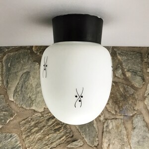 Op de afbeelding: Witte glazen plafondlamp met een zwarte metalen basis en een eenvoudig geometrisch ontwerp. De lamp heeft een afgeronde vorm en is versierd met zwarte lijnen en stippen.