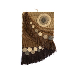 Op de afbeelding: Een geweven wandkleed in bruin en beige met een groot cirkelvormig ontwerp en franjes. De cirkel heeft een kleinere cirkel erin en is omgeven door kleinere cirkels.