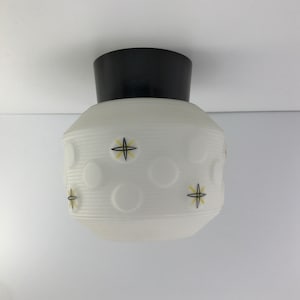 Puede incluir: Una lámpara de techo blanca con una base de metal negro. La pantalla de vidrio tiene una superficie texturizada con círculos en relieve y diseños de estrellas amarillas y negras.