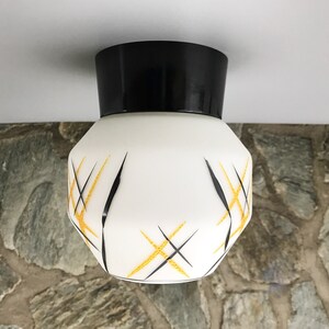 Può includere: Un apparecchio di illuminazione a soffitto bianco con una base nera e un motivo geometrico di linee gialle e nere.
