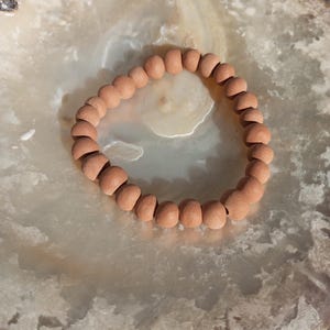 Könnte beinhalten: Ein Armband aus terrakottafarbenen Perlen. Das Armband besteht aus runden, gleichmäßig großen Perlen. Die Perlen sind zu einem Kreis aufgefädelt. Das Armband liegt auf einer hellen, strukturierten Oberfläche. Die Perlen haben einen Durchmesser von ca. 1 cm.