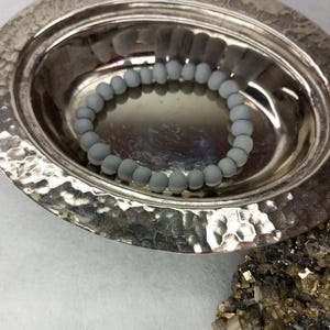 Può includere: Un piatto in metallo argentato martellato contiene un braccialetto di perline grigio chiaro. Il piatto è di forma ovale e poggia su una superficie bianca. Un gruppo di cristalli di pirite dorati si trova in basso a destra.