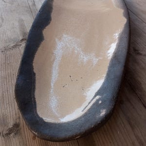 May include: An oval ceramic serving dish with a light tan interior and a dark grey, textured rim. The dish has a glossy finish and is placed on a wooden surface. The design is simple and rustic, suitable for serving food.