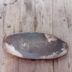 May include: A ceramic serving platter, oval in shape, with a mottled grey and white surface. The edges are uneven. The platter is displayed on a wooden surface.