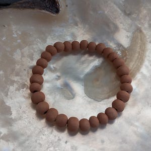 Könnte beinhalten: Ein armband aus terrakottafarbenen Perlen. Das Armband besteht aus runden, matten Perlen. Das Armband wird auf einer perlmuttartigen, strukturierten Oberfläche präsentiert, wodurch ein Kontrast zwischen den Erdtönen des Armbands und dem schillernden Hintergrund entsteht.