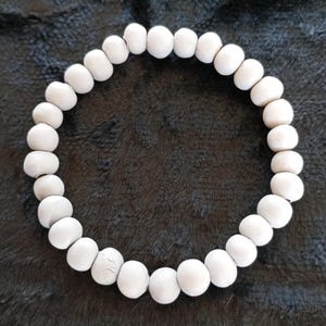 Könnte beinhalten: Ein weißes Perlenarmband. Das Armband besteht aus runden, hellen Perlen, die aneinandergereiht sind. Die Perlen scheinen aus einem matten Material zu bestehen. Das Armband wird auf einer dunklen, strukturierten Oberfläche gezeigt. Ca. 18 cm Umfang.