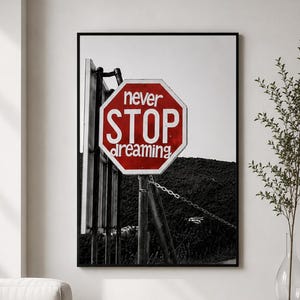 May include: A framed wall art print featuring a black and white photograph of a red octagonal stop sign. The sign has white text that reads never STOP dreaming. The sign is mounted on a metal pole with a chain attached, set against a dark landscape.