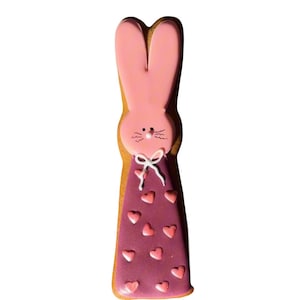May include: A pink bunny-shaped biscuit with a purple dress decorated with pink hearts. The bunny has long ears and a white bow. The biscuit is a light brown colour.