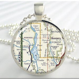 May include: A silver-toned pendant necklace featuring a circular map design. The map shows a detailed geographical layout with blue water features, green forests, and place names like Priest Lake and Kaniksu. The pendant hangs from a beaded chain.