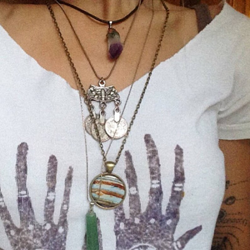 May include: A layered necklace with multiple pendants, including a purple crystal, a silver charm with a geometric design, and a round pendant with a striped pattern.