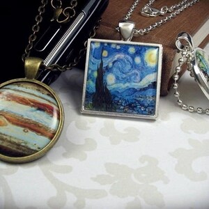 May include: Three necklaces with different pendants. One pendant features a painting of a starry night sky, another a painting of Jupiter, and the third a map. The necklaces are on a white surface.