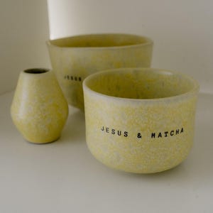 Könnte beinhalten: Ein Set aus drei gelben Keramikgefäßen mit gesprenkelter Oberfläche. Die vordere Schale trägt den schwarzen Schriftzug "JESUS & MATCHA" an der Seite, während die größere Schale dahinter das Wort "JESUS" in schwarzen Buchstaben auf der Außenseite zeigt.