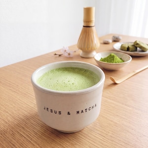 Könnte beinhalten: Eine hellfarbene Keramikschale gefüllt mit schaumigem grünem Matcha-Tee, mit dem schwarzen Schriftzug "JESUS & MATCHA" auf der Vorderseite. Daneben befinden sich ein Bambusbesen, eine kleine Schale mit Matcha-Pulver und ein Bambuslöffel.