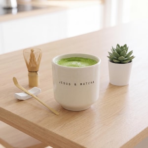 Könnte beinhalten: Ein weißer Keramikbecher gefüllt mit leuchtend grünem Matcha-Tee, der auf der Vorderseite den schwarzen Schriftzug 'JESUS & MATCHA' trägt. Neben dem Becher stehen ein heller Bambusbesen, ein Bambuslöffel auf einer weißen Ablage und eine kleine Sukkulente.