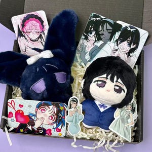 Può includere: Una scatola contiene un peluche a forma di coniglio blu scuro con dettagli viola, una bambola in abito blu e biglietti illustrati in stile anime. I biglietti presentano personaggi anime in varie pose.