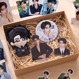 May include: A cardboard gift box filled with shredded paper containing a plush doll, a circular pin button with the text Dreamland, and two acrylic standees featuring portraits of a young man in a suit, surrounded by various photo cards.