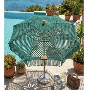 May include: A green macrame parasol with a wooden handle and fringe. The parasol is open, casting a shadow on the stone patio. A small woven handbag and glasses of water sit on a wooden table. The parasol has the text "RCT".