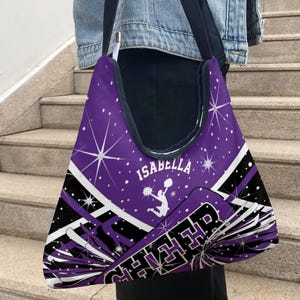 May include: A purple hobo bag featuring a white graphic of a cheerleader holding pom-poms, the name Isabella, and the word CHEER. The design includes white starbursts, diagonal black and white stripes, and a black strap against a purple background.