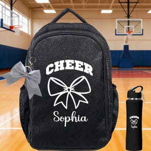 May include: Black glitter backpack with the word "CHEER" and a white bow graphic. The name "Sophia" is below the bow. A silver bow keychain hangs from the backpack. A black water bottle with matching design is also shown.