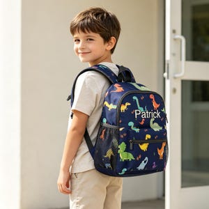 Puede incluir: Una mochila azul marino con un patrón de dinosaurios coloridos y el nombre Patrick impreso en blanco en el bolsillo frontal. La bolsa incluye correas negras ajustables para los hombros, un asa superior y bolsillos laterales de malla negra.
