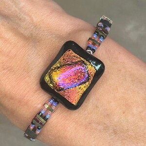 Puede incluir: Una pulsera con una cuenta central rectangular de vidrio negro con un centro iridiscente en tonos naranja, rosa y dorado. La banda está formada por dos filas paralelas de pequeñas cuentas de vidrio multicolor y espaciadores metálicos.