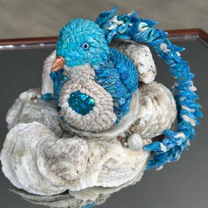 May include: A handcrafted sculpture of a blue bird with a copper beak, perched on a bed of seashells. The bird's body features intricate detailing and a shimmering, iridescent center. The sculpture is a unique piece of art.
