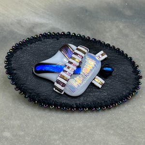 May include: An abstract glass art piece with a black leather base. The glass features white, blue, and iridescent accents, with a beaded border. The piece has a unique, artistic design.