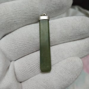Pode incluir: Um pingente retangular de jade verde-oliva, com uma tampa prateada e uma pequena argola para uma corrente. O pingente polido mostra variações naturais de cor. Adequado para colares, mede aproximadamente 4 cm de comprimento.