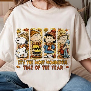 May include: A cream-colored t-shirt featuring a graphic of Snoopy, Charlie Brown, Lucy, and Linus wearing winter hats and scarves. The text "IT'S THE MOST WONDERFUL TIME OF THE YEAR" is printed in brown below the characters, surrounded by autumn leaves.