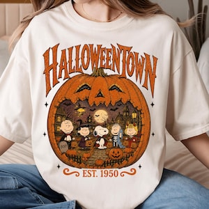 May include: An off-white t-shirt featuring a large orange pumpkin graphic with the text HALLOWEENTOWN and EST. 1950. Inside the pumpkin, Peanuts characters are dressed in Halloween costumes in a spooky town setting with bats and a full moon.
