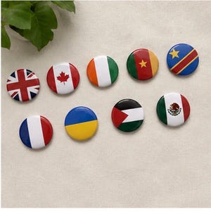 May include: Nine circular pin-back buttons featuring various national flags, including the United Kingdom, Canada, Ireland, Cameroon, Democratic Republic of the Congo, France, Ukraine, Palestine, and Mexico, arranged in two rows on a light surface.