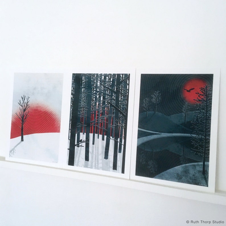 May include: Three framed prints with black and white illustrations of trees and a red sun. The first print shows a single tree with a red sun behind it. The second print shows a forest of trees with a red sun behind them. The third print shows a forest with a red sun and a black bird flying in the sky.