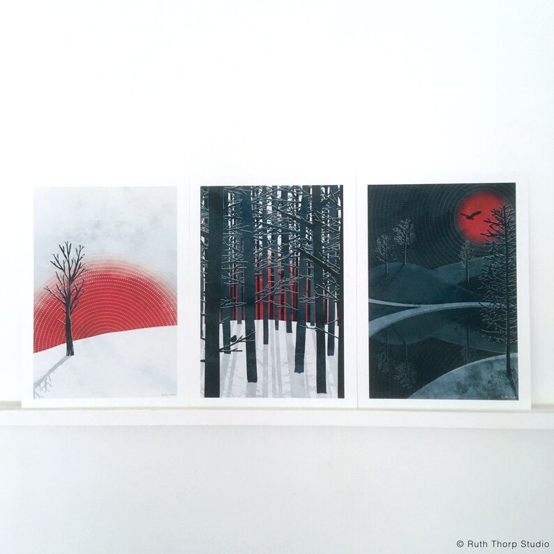 May include: Three framed prints with black and white forest scenes. The left print features a single tree in a snowy landscape with a red and white sunset. The middle print shows a forest with tall trees and a red and white sunset. The right print shows a dark forest with a black bird flying over a lake.