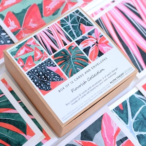 Könnte beinhalten: Ein Kartenset mit 12 Karten und Umschlägen aus der "Flourish Collection". Die Karten zeigen lebendige botanische Illustrationen in Rosa, Grün und Weiß. Die Box ist hellbraun mit einem weißen Etikett mit der Aufschrift "BOX OF 12 CARDS AND ENVELOPES."