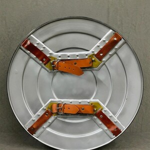 May include: A silver-colored, round shield with a riveted metal frame. It features two orange leather straps with buckles and brown leather accents. The shield has a cross-shaped design with yellow and brown details.