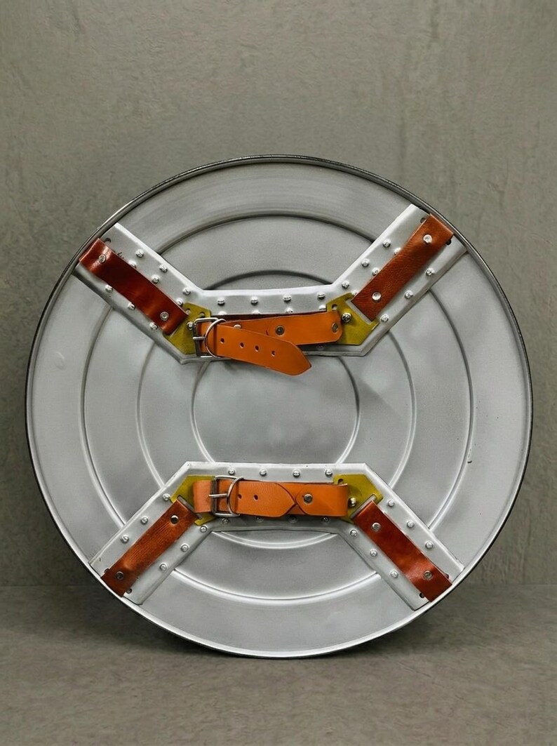 May include: A silver-colored, round shield with a riveted metal frame. It features two orange leather straps with buckles and brown leather accents. The shield has a cross-shaped design with yellow and brown details.