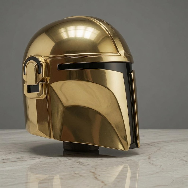 May include: A polished gold-colored helmet featuring a smooth, rounded dome and a T-shaped black visor. The side of the helmet displays a raised rectangular ear piece with black accents, and the overall design has a reflective, metallic finish.