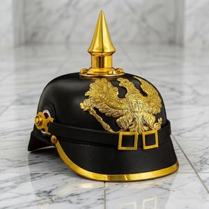 May include: Black and gold military helmet with a prominent gold eagle emblem and a pointed top. The helmet features gold trim and accents, including a chin strap with square buckles. The design is ornate and detailed, suggesting a historical or ceremonial purpose.