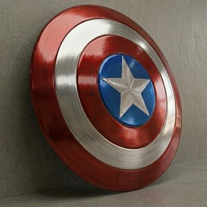 May include: A metallic red, silver, and blue shield with a star emblem. The shield has a circular design with concentric rings of red, silver, and blue. A silver star is centered on the blue circle. The shield is leaning against a gray surface.