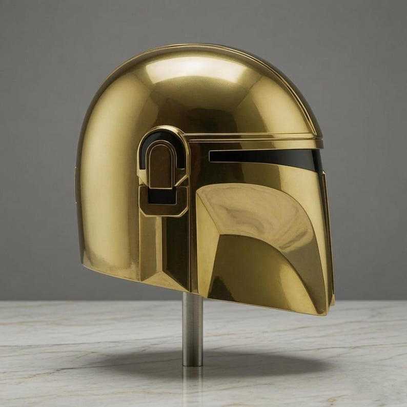 May include: A polished gold-colored helmet featuring a smooth, rounded dome, a T-shaped visor opening with a black horizontal band, and geometric side detailing. The helmet is mounted on a vertical silver-toned metal stand for display purposes.