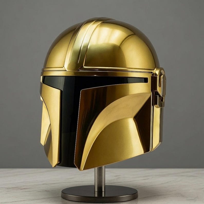 May include: A polished gold-colored helmet featuring a smooth, rounded dome, a prominent central ridge, and a wide, dark, horizontal visor section. The helmet is mounted on a simple, circular metal stand with a vertical support post.